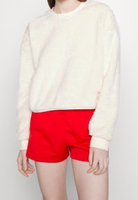 Vit, fluffig långärmad sweatshirt kombinerad med röda shorts. Sweatshirten har en rund halsringning och ribbade muddar.