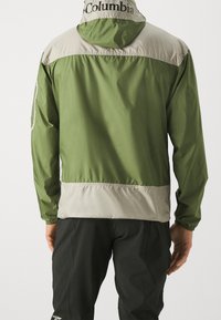 Chaqueta de exterior verde y beige con capucha ajustable, puños elásticos y un logo en la parte trasera. Textura suave, diseño ligero.