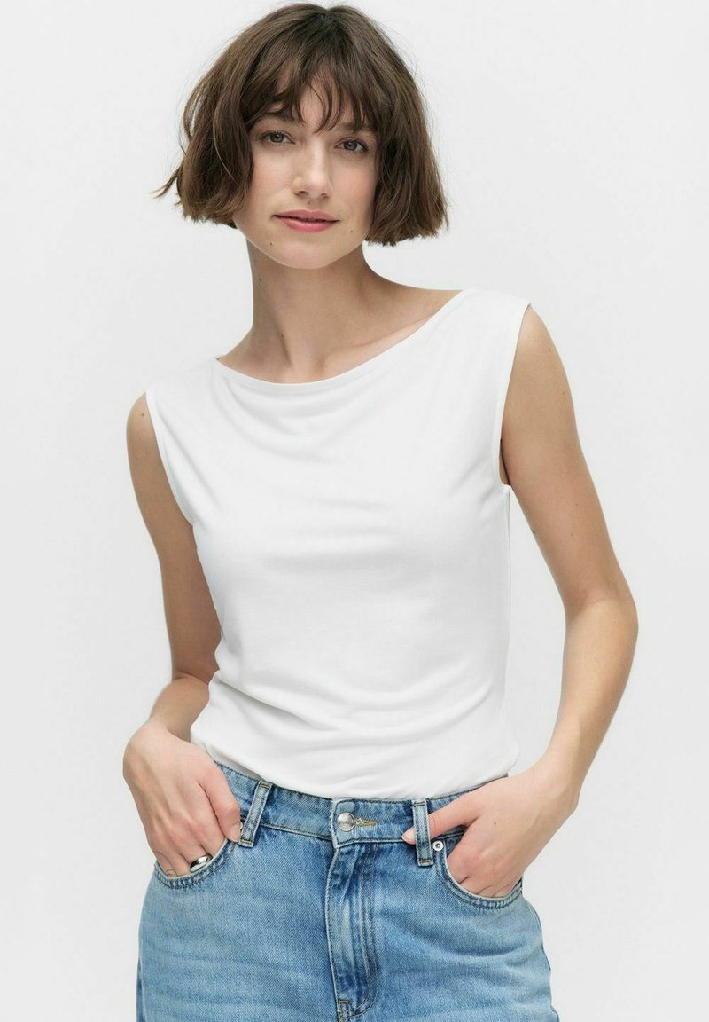 Haut sans manches blanc en tissu doux, avec un décolleté drapé et une coupe décontractée, associé à un jean en denim bleu.