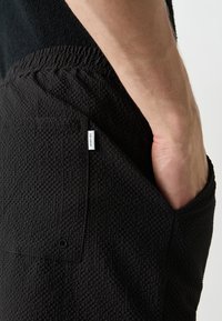 Gros plan d'une personne portant un short noir texturé avec une étiquette blanche "LES DEUX", la main partiellement dans la poche latérale.
