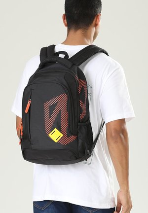 Homme portant un t-shirt blanc et un jean bleu, portant un sac à dos noir avec un motif de rayure diagonale rouge et une étiquette jaune.