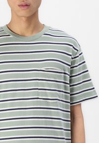 Camiseta de manga corta a rayas en verde, blanco y azul marino, con un bolsillo en el pecho, cuello redondo y textura de algodón suave.