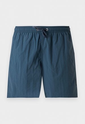 Homme musclé debout pieds nus portant un short de bain bleu foncé, bras croisés, sur un fond blanc uni.