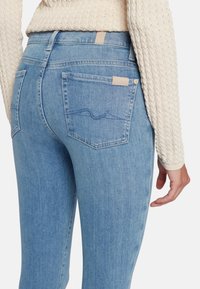 Jeans in denim azzurro chiaro con vestibilità slim, due tasche posteriori e un delicato dettaglio di cucitura ondulata. Il materiale appare liscio con leggera texture.