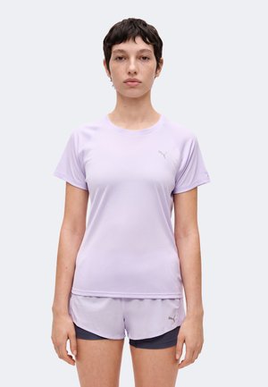 Ung person med kort mørkt hår iført lys lilla sportstrøje og matchende shorts, stående mod en ensfarvet baggrund.