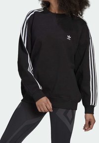 Svart sweatshirt med rund halsringning, vit tredelad design på ärmarna och liten vit logotyp. Kombinerad med texturerade svarta mönstrade leggings.