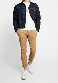 Veste en jean foncé portée sur un t-shirt blanc avec un logo en forme de bouclier, associée à un pantalon chino kaki et des baskets blanches, se tenant devant un fond uni.