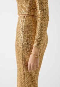 Goldfarbenes zweiteiliges Paillettenoutfit mit eng anliegenden langen Ärmeln und einem hoch taillierten Rock mit einem Reißverschluss auf der Rückseite. Kombiniert mit einem goldenen Armband.