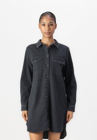 Black denim shirt dress with long sleeves, front button closure, two chest pockets, and a curved hemline. Textured fabric with a casual fit.