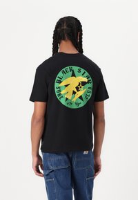Black t-shirt with short sleeves, featuring a vibrant green circular graphic of an eagle and text, worn with blue denim pants.