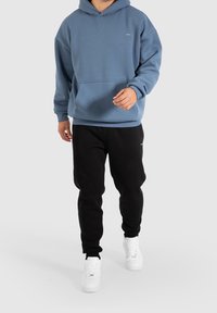 Felpa con cappuccio blu dotata di tasca frontale, abbinata a pantaloni della tuta neri e sneaker bianche. Fit casual e rilassato con un design semplice.