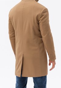 Ljusbrun skräddarsydd kappa med en strukturerad yta, bakre slits och tre knappar på varje ärm, bärs över blå jeans.