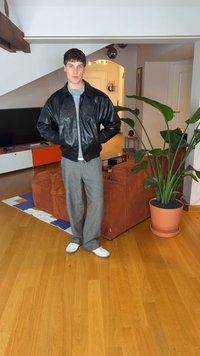 Schwarze Leder-Bomberjacke, graue Wollhose und weiße Sneakers. Innenraum mit einem Topfpflanze und einem Sofa im Hintergrund.