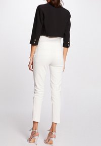 Woman wearing white high-waisted pants, black cropped blazer with gold buttons, and silver high-heeled sandals, standing against plain background.