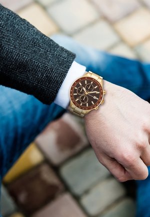 Goldfarbene Uhr mit braunem Zifferblatt, drei Unterzifferblättern, Datumsanzeige und Metallarmband. Am Handgelenk mit einem Fischgrät-Manschettenaufschlag getragen.