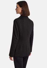 Kobieta z prostymi brązowymi włosami nosi dopasowaną czarną marynarkę i czarne spodnie, stoi bokiem na jednolitym białym tle.