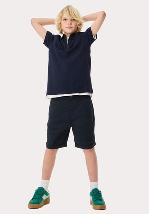 Marineblauwe rits polo, witte onderhemd, donkere shorts en groene sneakers met een textuur op de zolen. Ruime pasvorm en opgerolde shorts.