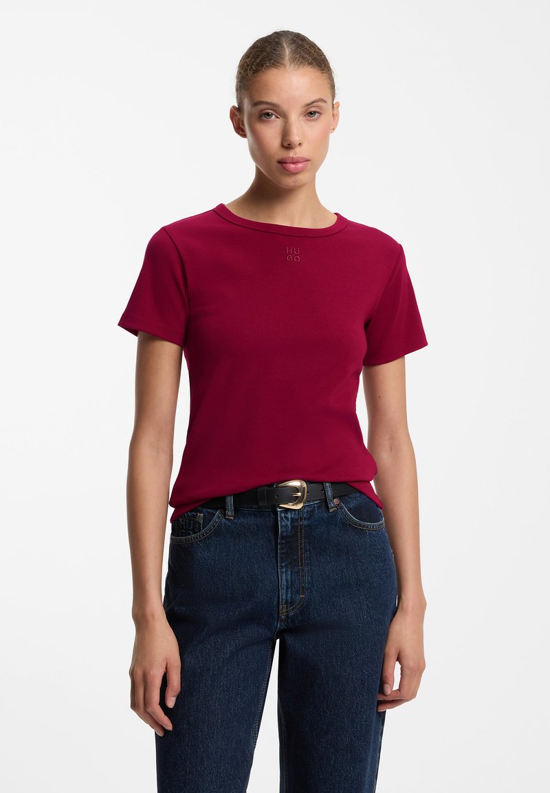 Femme portant un t-shirt ajusté rouge foncé à manches courtes et un jean bleu foncé avec une ceinture noire, debout devant un fond clair uni.