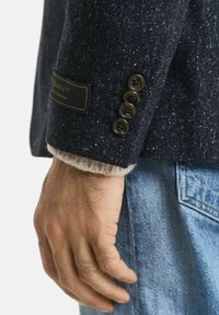 La mano di un uomo accanto alla manica di un blazer lungo di un blu scuro macchiato con quattro bottoni e un'etichetta GANT ben visibile, indossato sopra un maglione beige e jeans blu.