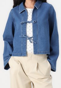 Veste en jean bleu au design raccourci, avec un col et des fermetures à nouer avec un ruban. Portée sur un haut blanc à pois et un pantalon beige.