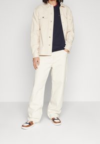 Giacca beige a bottoni sopra una t-shirt blu navy, abbinata a pantaloni bianchi a gamba larga e sneakers con dettagli marroni. Sfondo semplice.