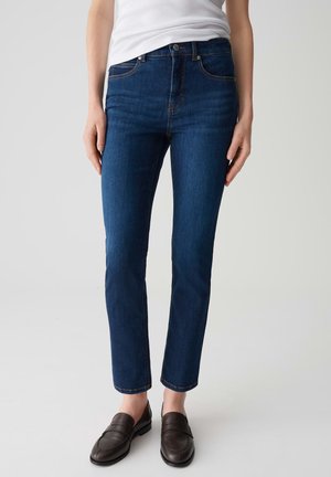 Jeans slim-fit blu scuro realizzati in denim, con un classico design a cinque tasche e dettagli di cuciture arancioni. Indossati con mocassini marroni scuri.