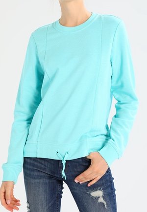 Personne portant un sweat-shirt à manches longues bleu clair avec un ourlet à cordon et un jean foncé déchiré, main posée sur la hanche.