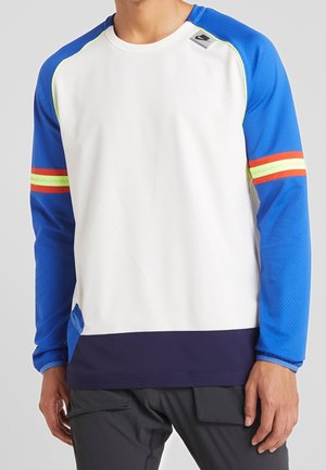Man draagt een sportshirt met lange mouwen met een witte voorkant, blauwe mouwen, rode en gele strepen op de armen en een donkerblauwe zoom.