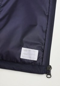 Námořnická modrá taštička na zip vyrobená z hladké nylonové tkaniny. Obsahuje bílou štítek s řádky pro údaje "JMÉNO" a "TELEFON" a kovové táhlo zipu.