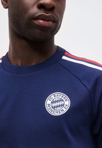 Άνδρας που φοράει μπλε σκούρο φούτερ FC Bayern München με κόκκινες και λευκές ρίγες στους ώμους, δείχνει το κάτω μέρος του προσώπου και τον λαιμό.