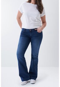 Femme portant un t-shirt blanc à manches courtes, un jean évasé bleu et des baskets blanches, debout, une main dans la poche, devant un fond uni.