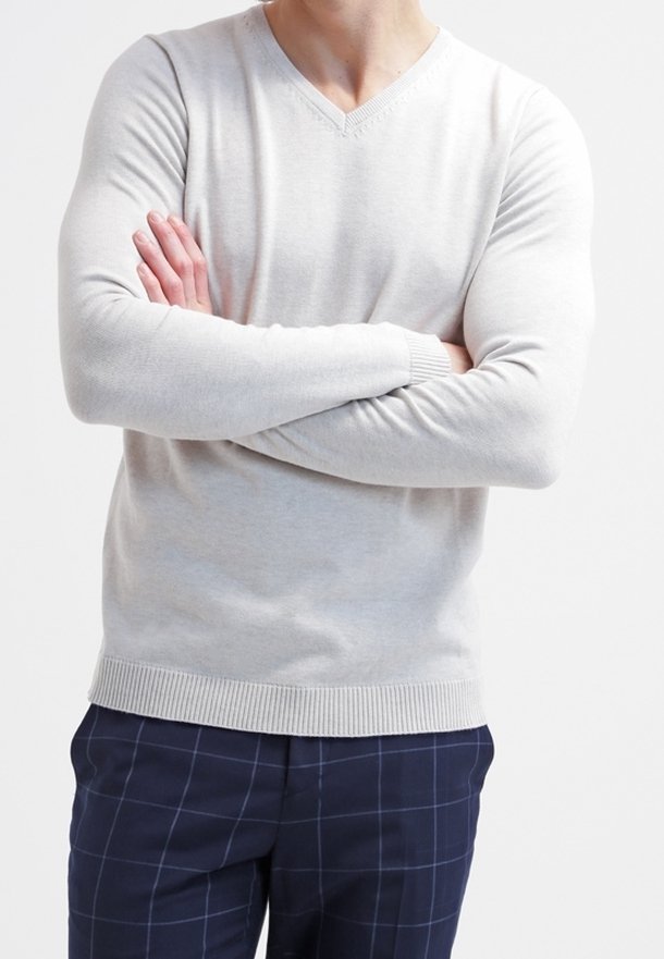 Personne portant un pull à col en V gris clair, les bras croisés, associé à un pantalon à carreaux bleu foncé, sur un fond blanc uni.