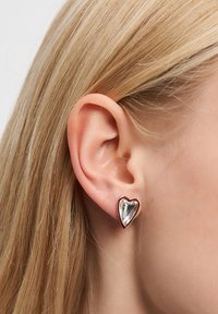 Herzförmige Ohrringe mit einem klaren Edelstein in der Mitte, umrahmt von einer rosevergoldeten Metallkante, die ein poliertes Finish und eine glatte Textur aufweisen.