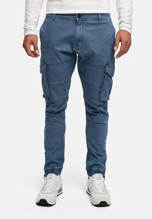 Pantalons cargo bleus en tissu mélangé de coton, dotés de plusieurs poches, de jambes fuselées et de poignets élastiques. Associés à des baskets blanches.