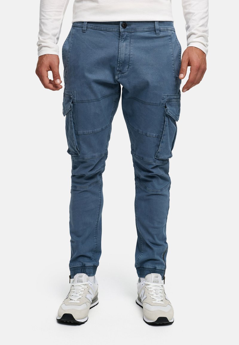 Blaue Cargo-Hosen aus einem Baumwoll-Mischgewebe, mit mehreren Taschen, schmal zulaufenden Beinen und elastischen Bündchen. Kombiniert mit weißen Sneakers.