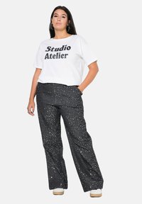 Camiseta blanca con estampado negro "Studio Atelier", combinada con pantalones amplios negros con un patrón salpicado en blanco. Zapatillas blancas completan el look.