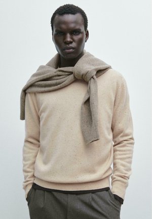Pull léger beige à finition texturée, associé à une écharpe en tricot marron drapée sur les épaules ; un pantalon gris rayé est visible.