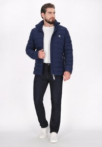Marinblå vadderad jacka med hög krage och dragkedjas stängning, prydd med en logotyp på bröstet, i kombination med mörka jeans och vita sneakers.