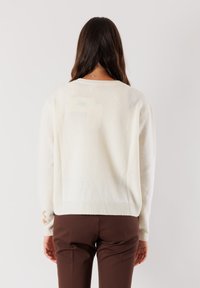 Donna con lunghi capelli castani che indossa un maglione crema e pantaloni marroni, in piedi di fronte a uno sfondo chiaro e semplice.