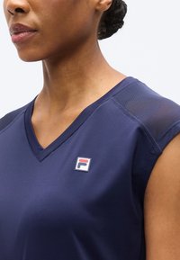 Μπλε ναυτικό αθλητικό τοπ με λαιμόκοψη σε σχήμα V, κοντά μανίκια και μικρό λογότυπο στο στήθος. Ομαλό ύφασμα με αντίθετα διακοσμητικά στους ώμους.