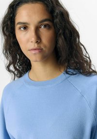 Sweatshirt en tricot bleu clair avec un col rond, des manches raglan et une texture douce. Vue en gros plan du col et de la partie supérieure du corps.