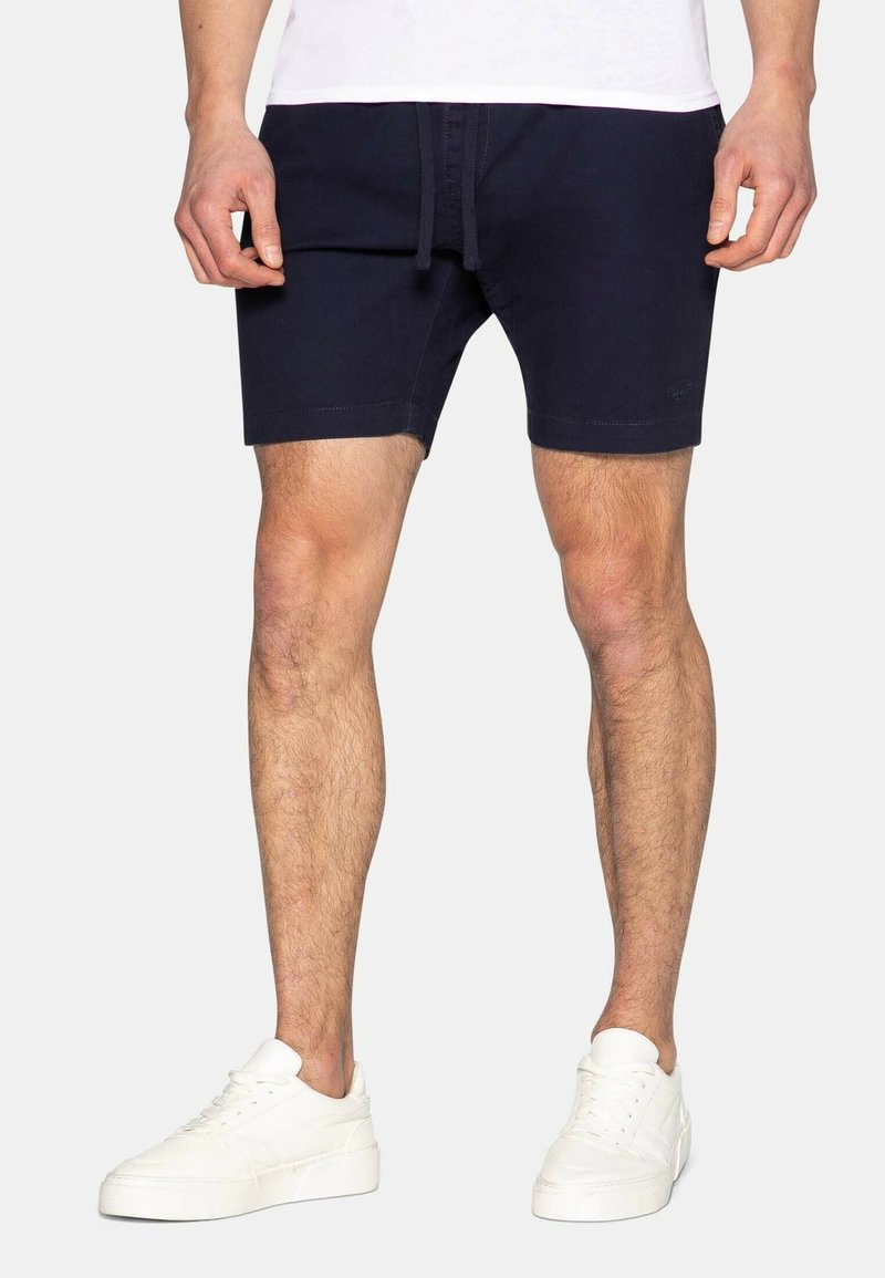 Homme portant un short noir et des baskets blanches, debout devant un fond clair uni.