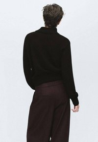 Persona con capelli corti e ondulati che indossa una maglietta nera a maniche lunghe e pantaloni larghi scuri, vista di spalle su uno sfondo semplice.
