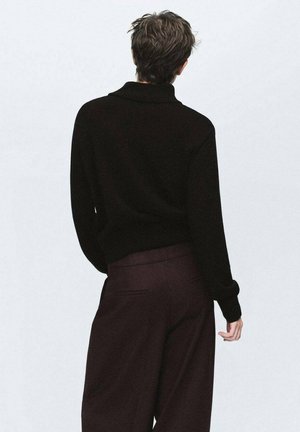 Persona con cabello corto y ondulado, vistiendo una blusa de manga larga negra y pantalones oscuros de pierna ancha, vista desde atrás contra un fondo liso.