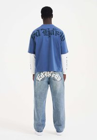 Blaues T-Shirt mit kurzen Ärmeln und großen schwarzen Buchstaben auf dem Rücken, getragen über einem weißen Langarmshirt, kombiniert mit hellblauen, weiten Jeans mit weißen Logoakzenten.