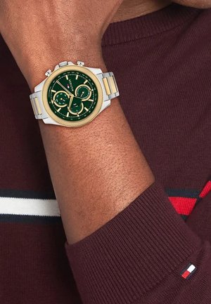 Chronograf s zeleným ciferníkom a zlatými akcentmi, s strieborným nerezovým puzdrom, predvedený na zápästí v bordo svetri.