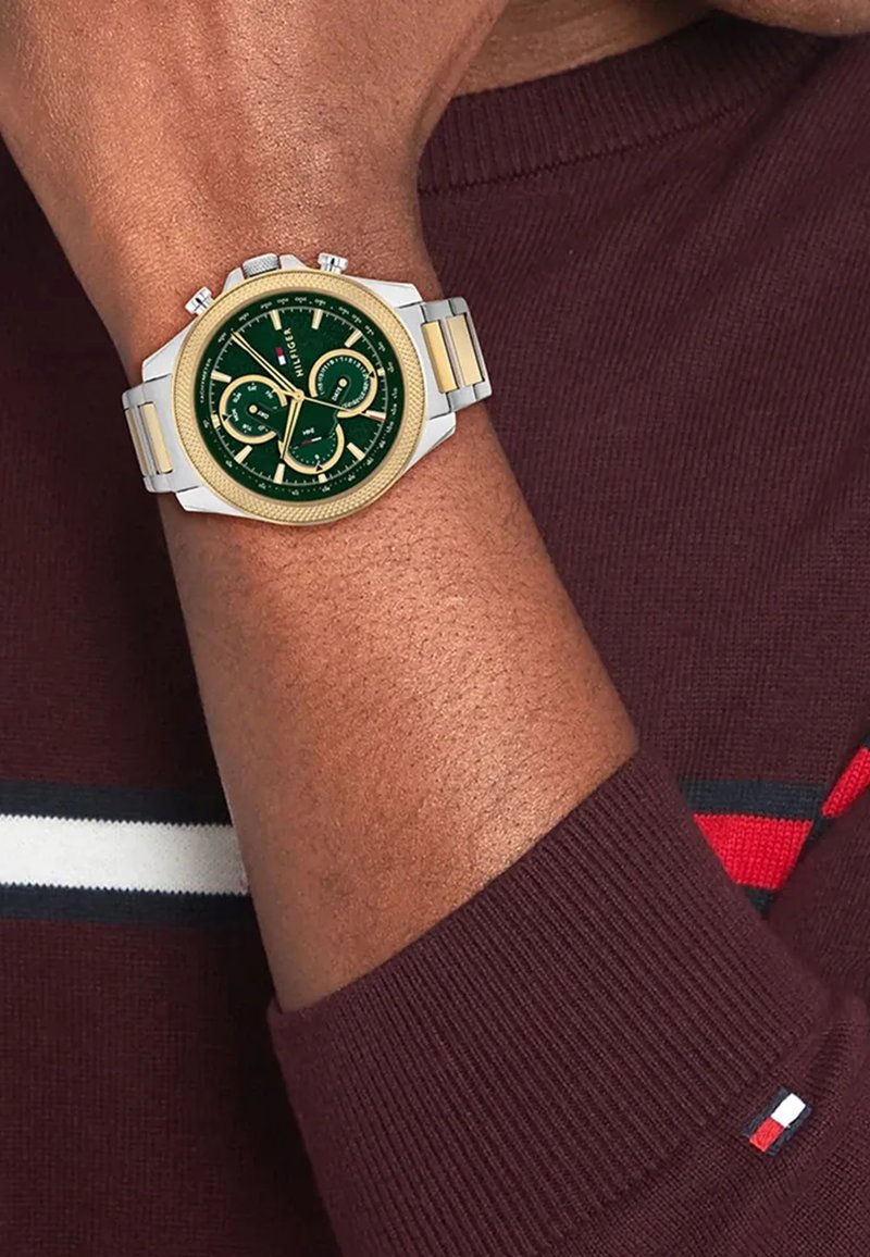 Grüne Zifferblatt-Chronographuhr mit goldenen Akzenten und silbernem Edelstahlgehäuse, am Handgelenk mit einem kastanienbraunen Pullover präsentiert.