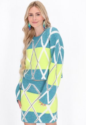 Kvinde med langt blondt hår iført en blå, gul og hvid mønstret sweater og matchende nederdel, smilende mod en hvid baggrund.