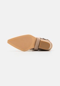 Zapato de tacón alto beige con punta puntiaguda, parte superior de ante y una correa en el tobillo. La suela tiene textura para mejor agarre y presenta una forma cónica.