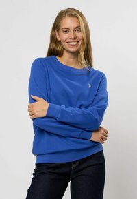 Felpa blu a girocollo realizzata in tessuto morbido, con polsini e bordo a costine, e un piccolo logo sul petto sinistro. Indossata con jeans scuri.